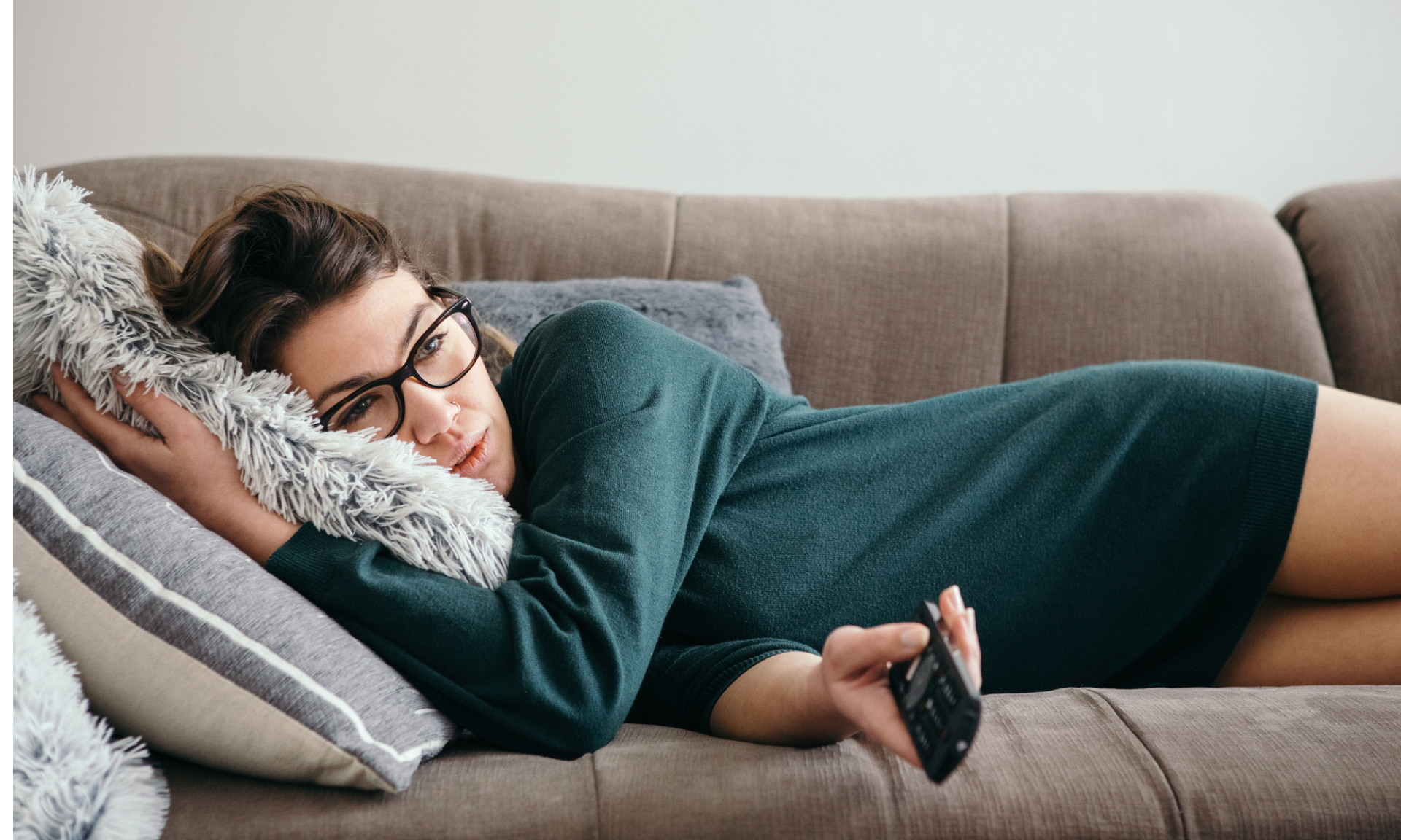 A woman laying on the couch holding the remote toward the TV