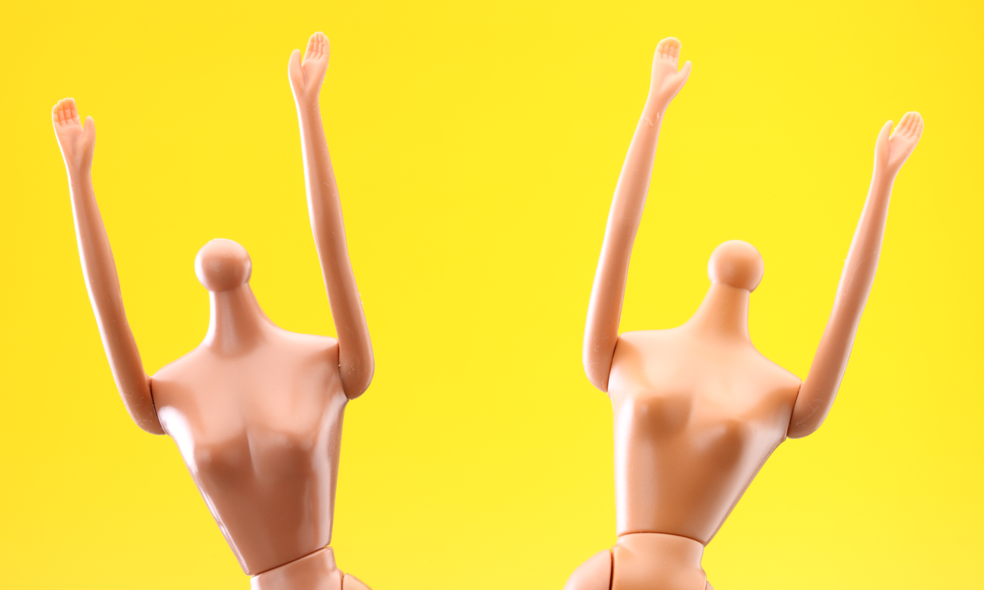 Two headless Barbie dolls with their arms in the air on a bright yellow backdrop