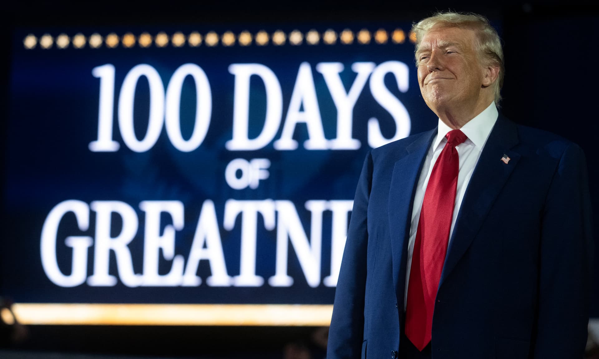Trump in front of a screen that says "100 days of greatness"